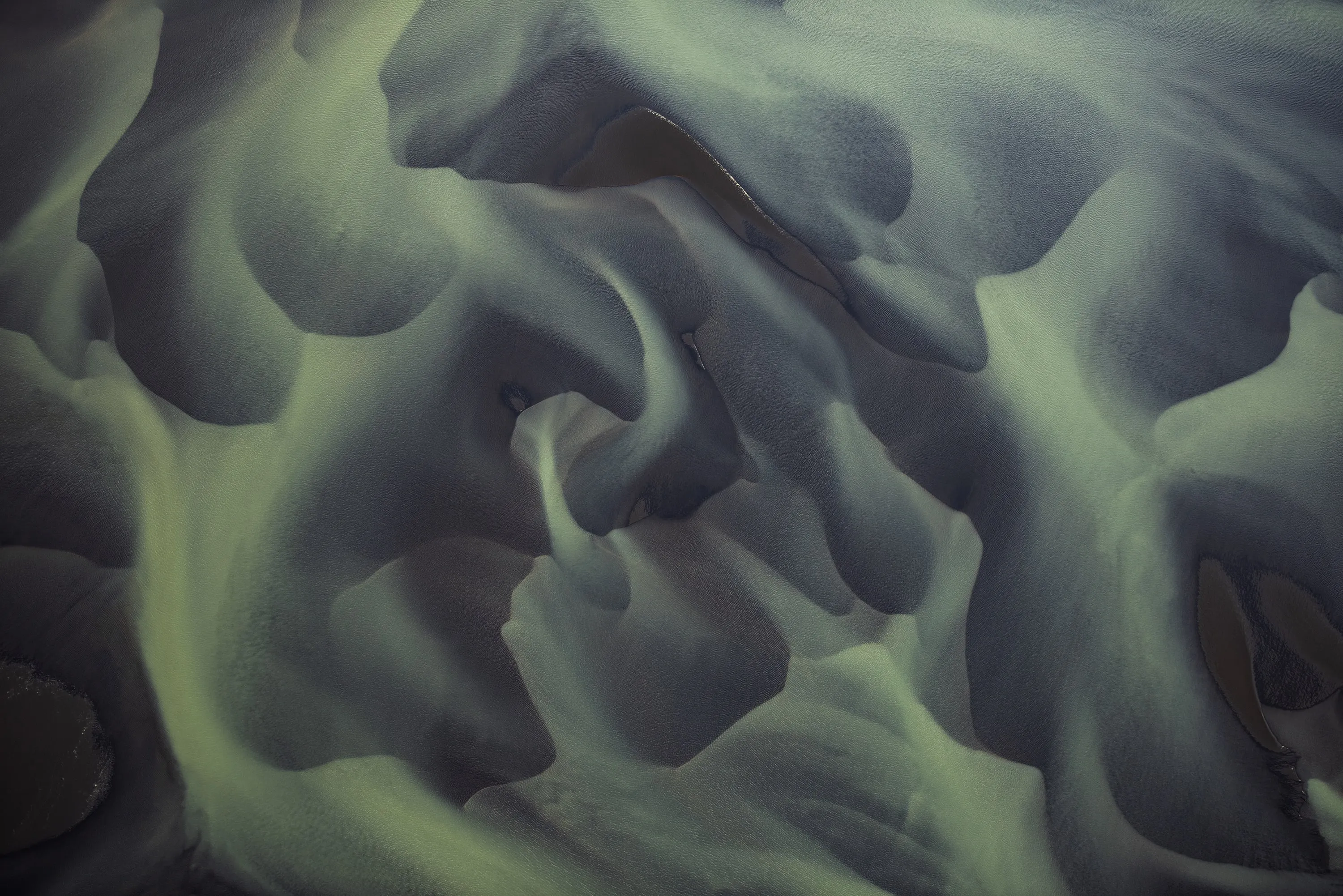 Aerial view of a braided river with dark water channels winding through light green sediment patterns.