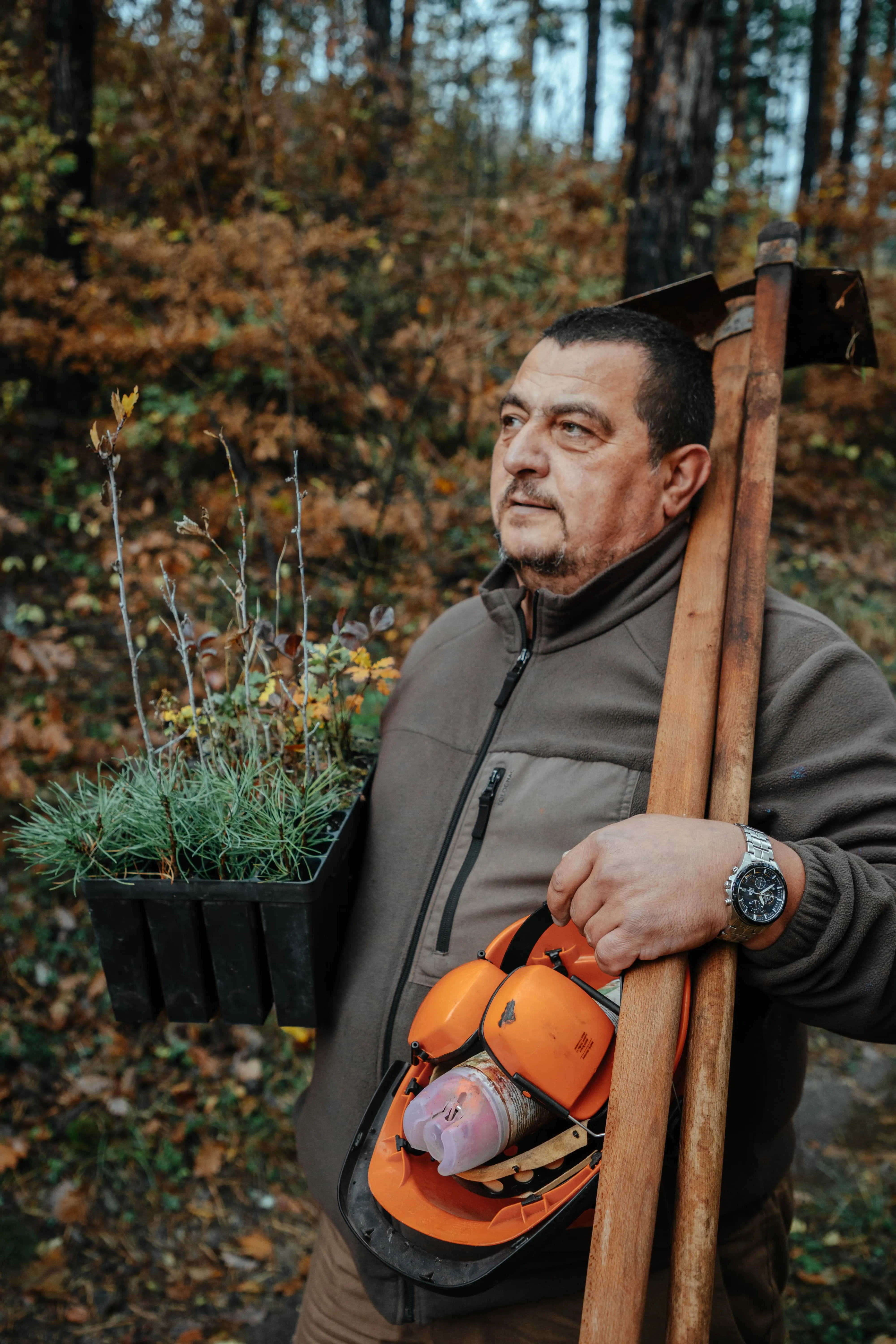 A person in outdoor clothing carrying tree saplings and equipment in a forest setting.