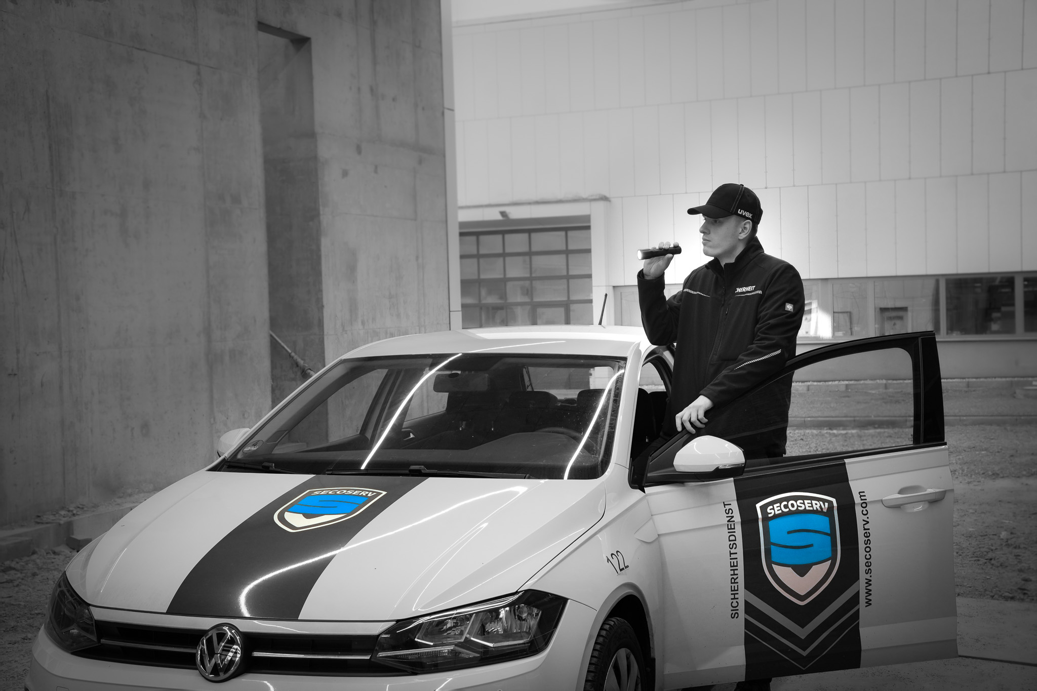 Sicherheitsdienst-Mitarbeiter mit Taschenlampe steht neben einem weißen SecoServ-Fahrzeug mit offener Tür.