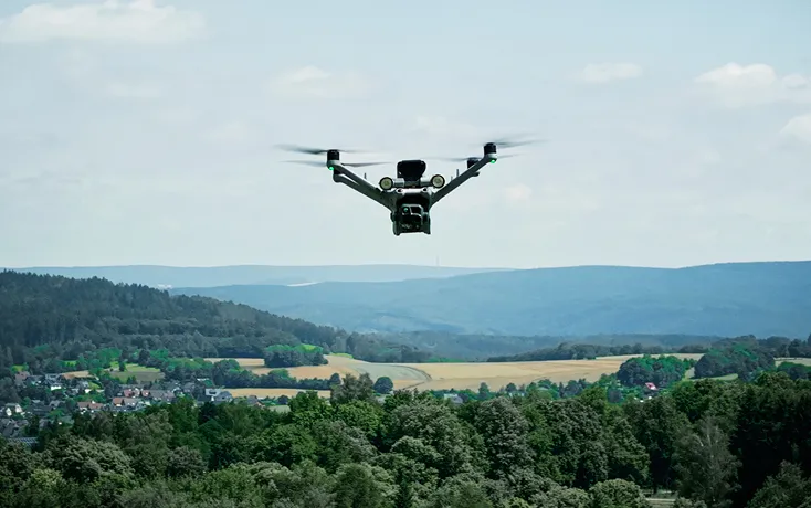 Drohne fliegt über bewaldete Landschaft mit Häusern und Hügeln im Hintergrund unter bewölktem Himmel.