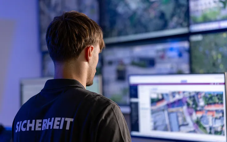 Ein Mann mit einem Hemd mit der Aufschrift "SICHERHEIT" auf dem Rücken schaut auf mehrere Monitore, die Karten und Luftaufnahmen anzeigen.