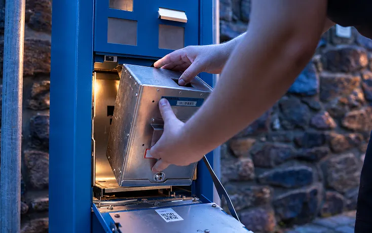 Eine Person entnimmt eine metallene Komponente aus einem blauen Automaten mit offener Tür, vor einer Wand aus unregelmäßigen Steinen.