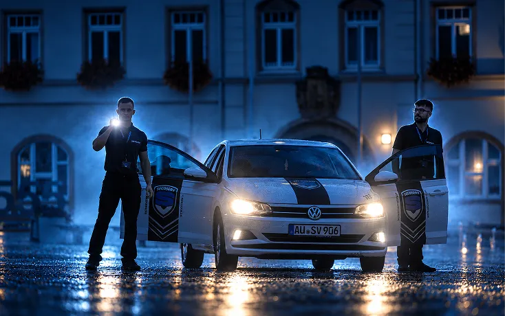 Zwei Sicherheitsmitarbeiter in dunkler Kleidung stehen nachts bei einem weißen Volkswagen mit geöffneter Fahrertür und Beifahrertür, die beide Sicherheitsschilder zeigen; einer hält eine Taschenlampe, und das Auto hat eingeschaltete Scheinwerfer auf nassem Kopfsteinpflaster vor einem beleuchteten Gebäude.