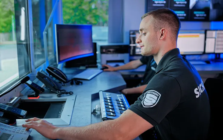 Ein Wachmann in Uniform sitzt an einem Kontrollraum mit mehreren Telefonen und Computermonitoren und bedient ein Bedienfeld.