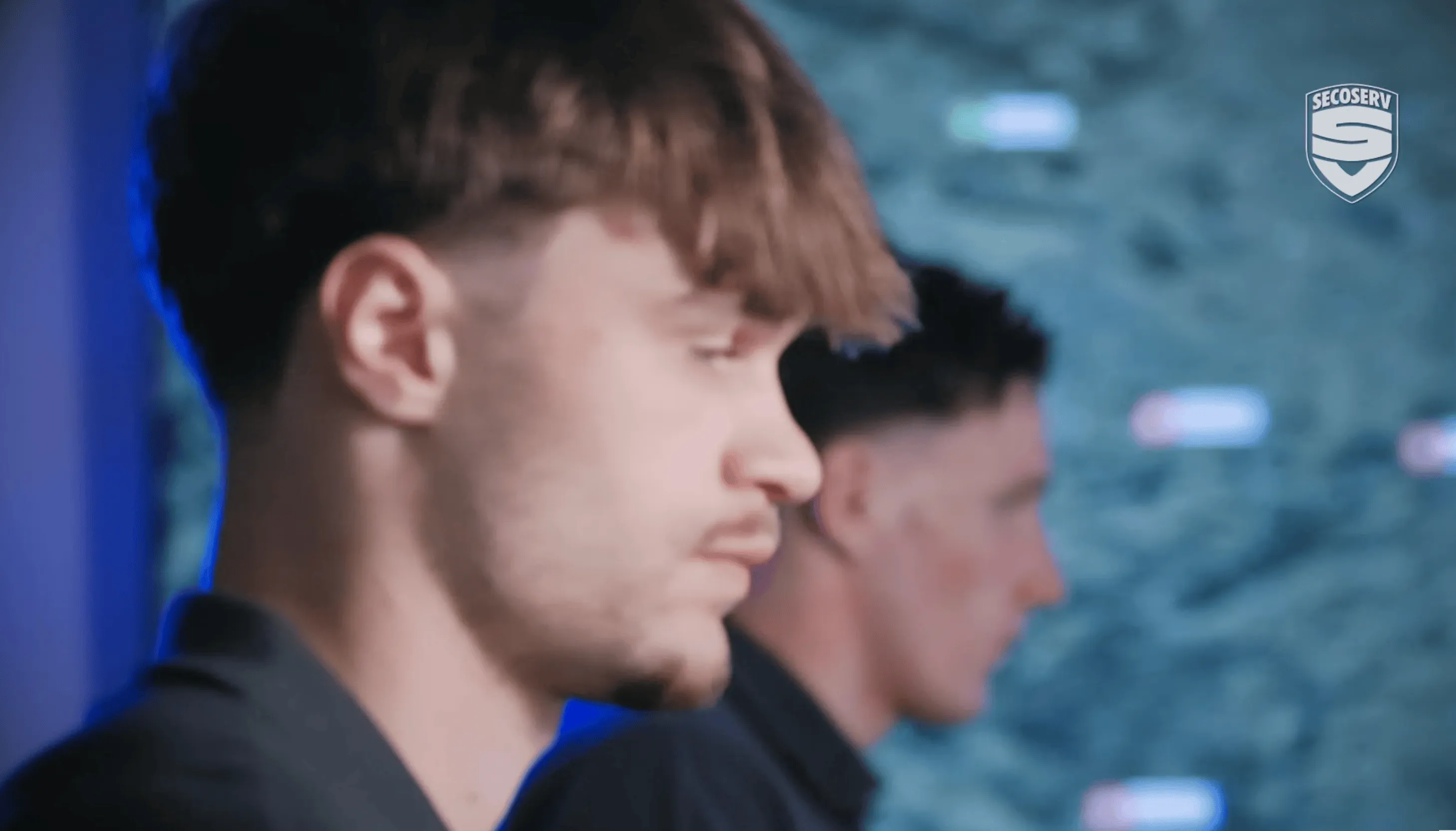 Seitenansicht von zwei jungen Männern mit kurzen Haaren, die ernst blicken, vor einem unscharfen blauen Hintergrund mit einem SECOSERV-Logo in der oberen rechten Ecke.