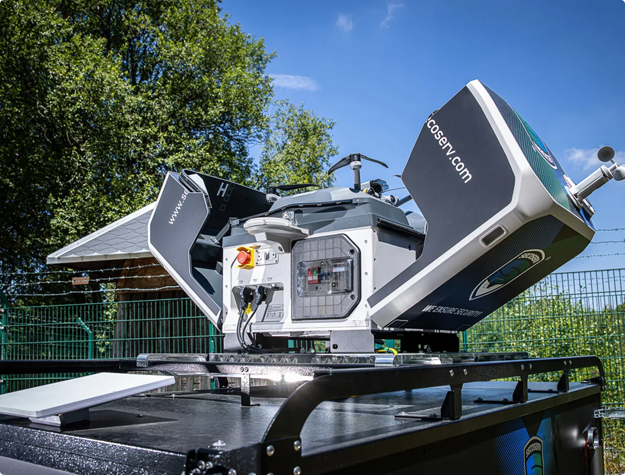 Ein modernes, weißes und schwarzes technisches Gerät mit geöffneten Seitenteilen, das auf einer schwarzen Plattform mit Metallstreben montiert ist, vor einem grünen Zaun und Bäumen unter blauem Himmel.