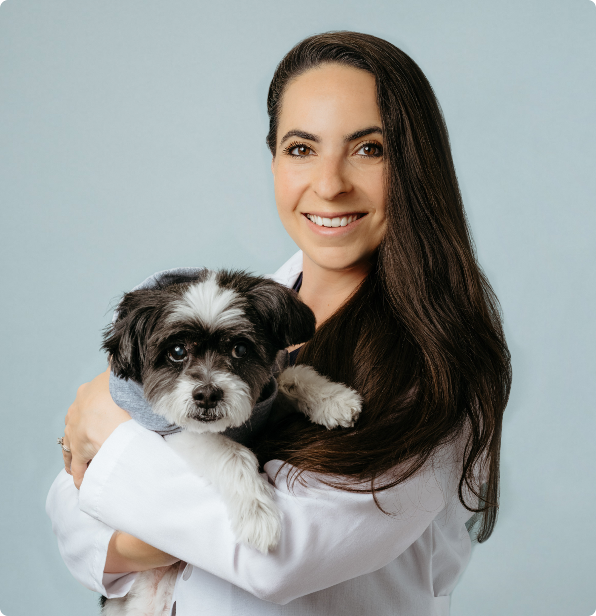 A woman in a surgical apron, wearing a ring, holds a small dog with a white coat and brown eyes, both smiling and looking at the camera, against a light blue background.
