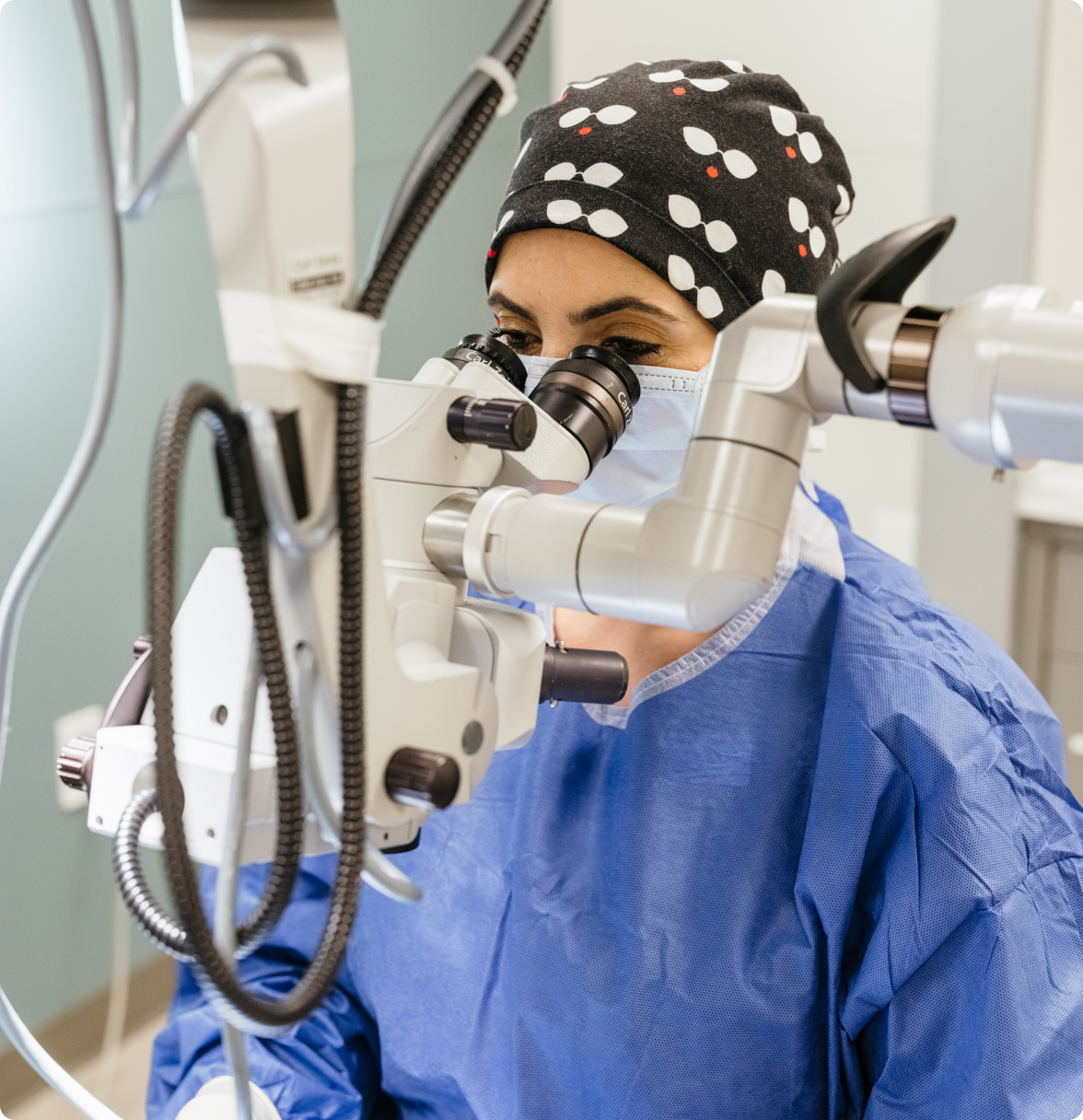 A modern operating room features a large white table with a black monitor, a black and silver chair, a white ceiling fan, a window with a green curtain, a white and black surgical table, and a white and black surgical chair, all set against a blue wall and wooden floor.