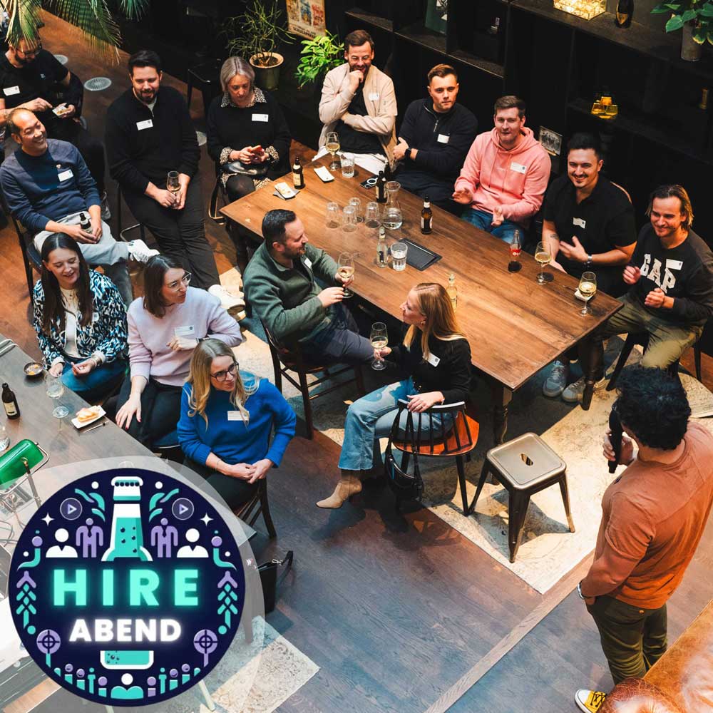 Eventteilnehmende des Hire Abends sitzen an einem langen Holztisch und hören dem Vortragenden zu.