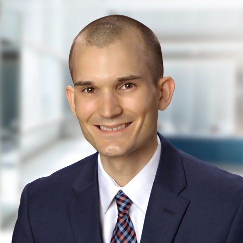 Smiling man with short hair wearing a dark suit, white shirt, and patterned tie in a bright indoor setting.