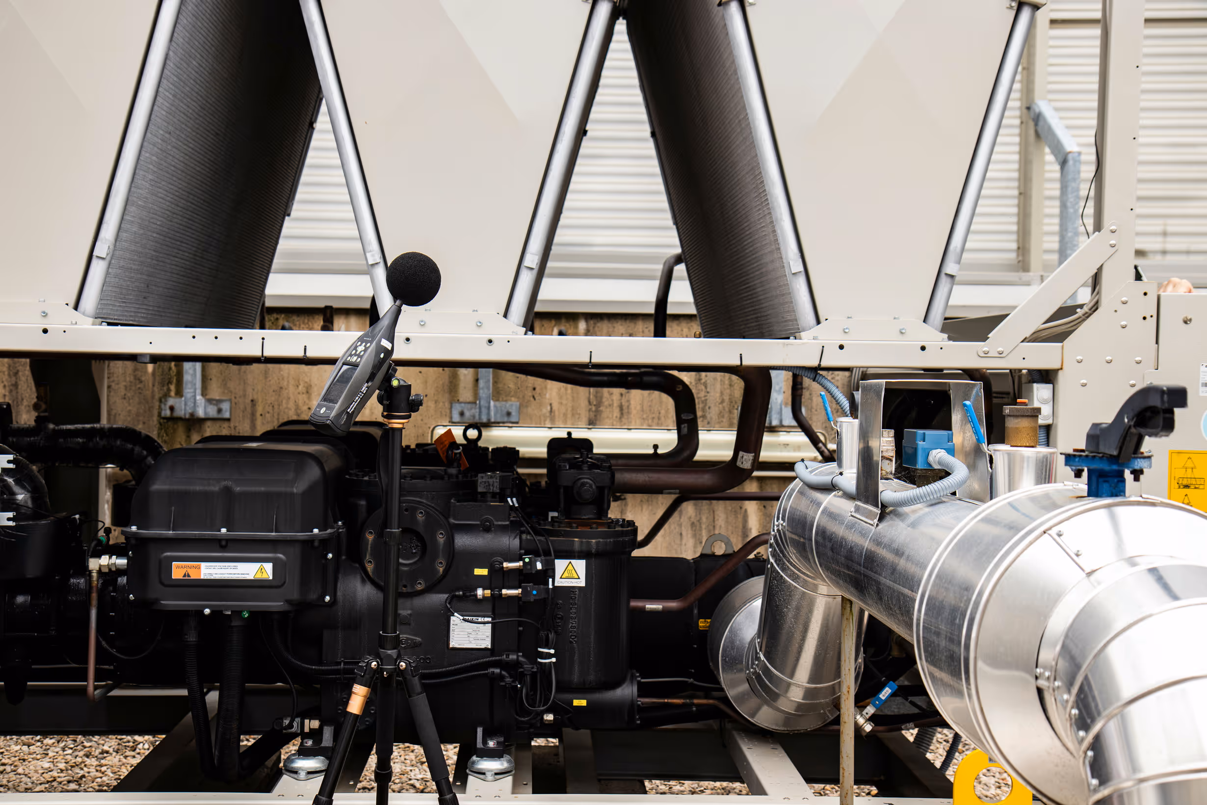 Industrial HVAC system with large metal pipes and a sound level meter on a tripod.