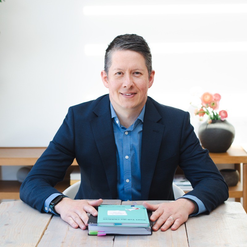 Man in a blue suit jacket sitting at a wooden table with a stack of books and notebooks in front of him, smiling at the camera.