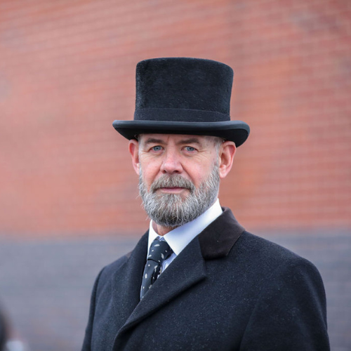 Middle-aged man with gray beard and mustache wearing a black top hat, black coat, white shirt, and polka dot tie standing outdoors against a brick wall.