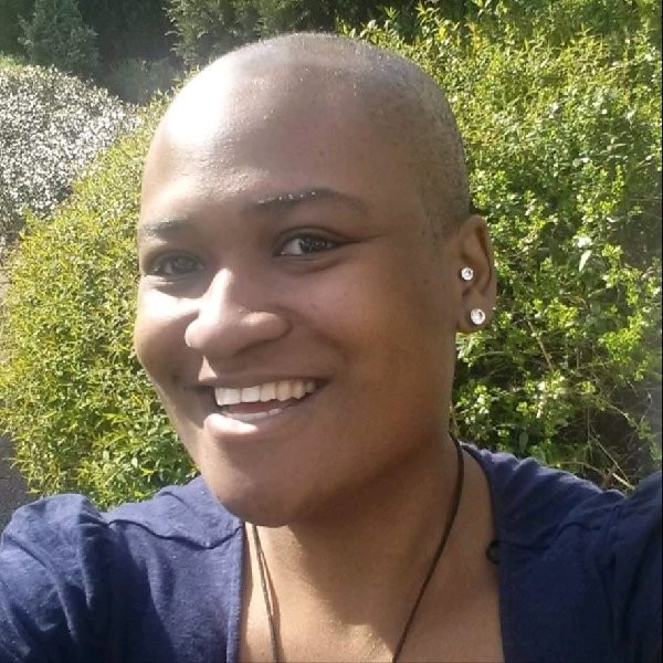 Smiling person with a shaved head wearing a dark top, standing outdoors with green foliage in the background.