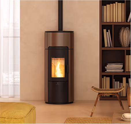 A modern cylindrical wood stove with black and brown finish, emitting a visible flame inside, positioned against a beige wall next to a bookshelf filled with books and decorative items, with a woven chair and a yellow cushion nearby.