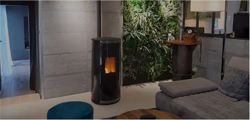 Modern interior living room with a gray sofa, small blue round ottoman, wooden coffee table, black cylindrical stove with fire, and a wall covered with lush green plants.