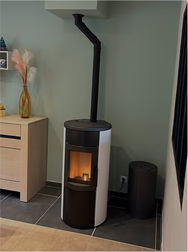 Modern black and white cylindrical wood-burning stove with a fire inside, installed against a green wall in a corner of a room.