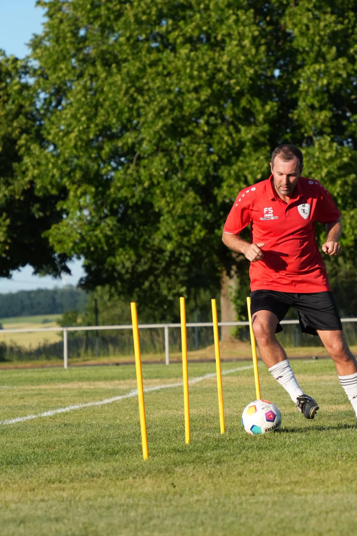 Ein Fußballspieler in rotem Trikot und Shorts dribbelt um aufgestellte Trainingshütchen auf einem grünen Fußballfeld unter einem strahlend blauen Himmel.