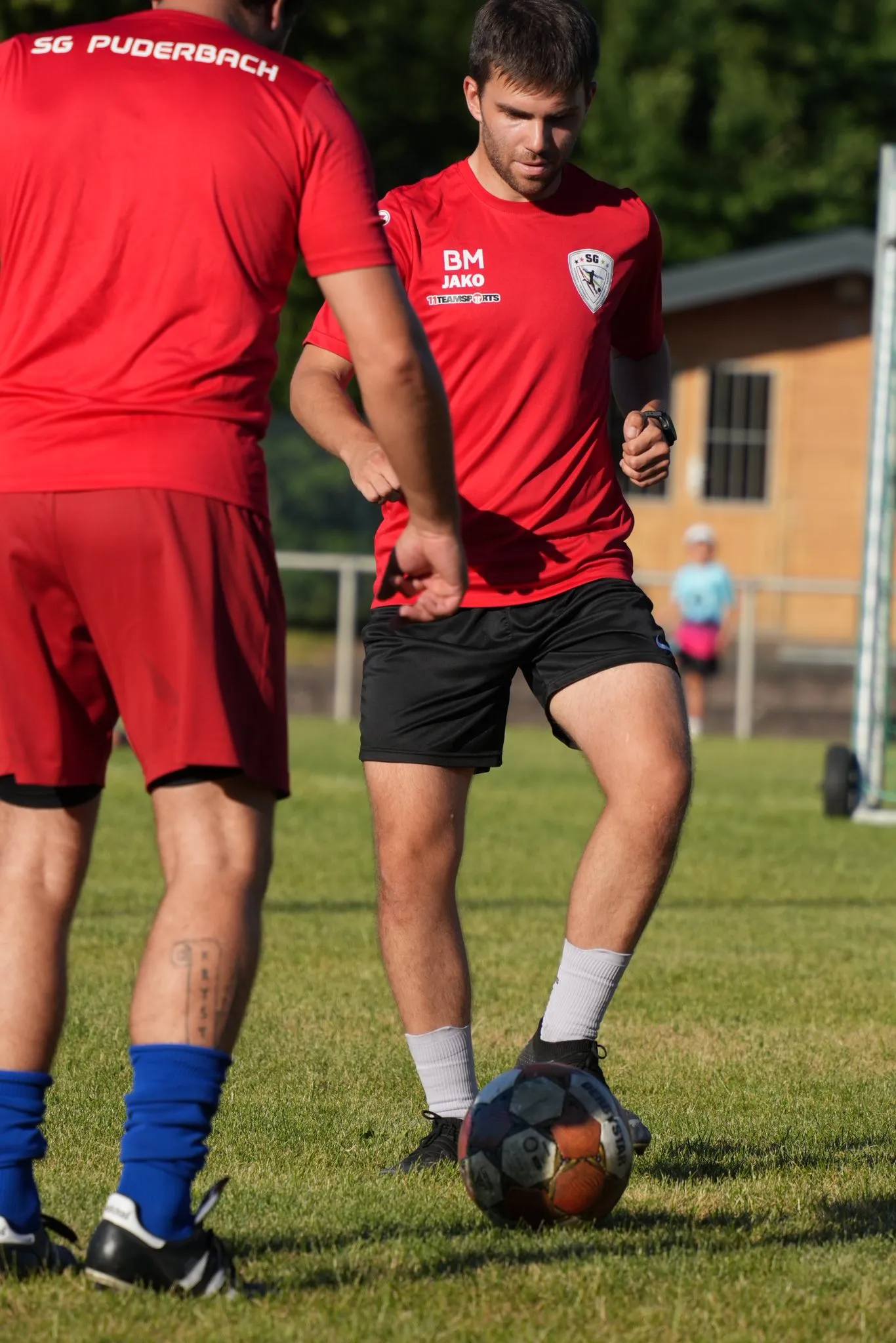Zwei Fußballspieler in roten Trikots üben auf einem grünen Rasenplatz. Ein Spieler dribbelt den Ball, während der andere ihn beobachtet. Im Hintergrund sind weitere Personen und ein Tor zu sehen.