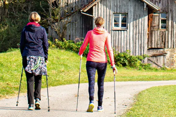 Zwei Frauen gehen mit Wanderstöcken auf einem gewundenen Weg, vorbei an einem alten, hölzernen Gebäude. Die erste Frau trägt eine dunkelblaue Jacke mit einem bunten Rock, während die zweite in einer roten Jacke und Leggings gekleidet ist.