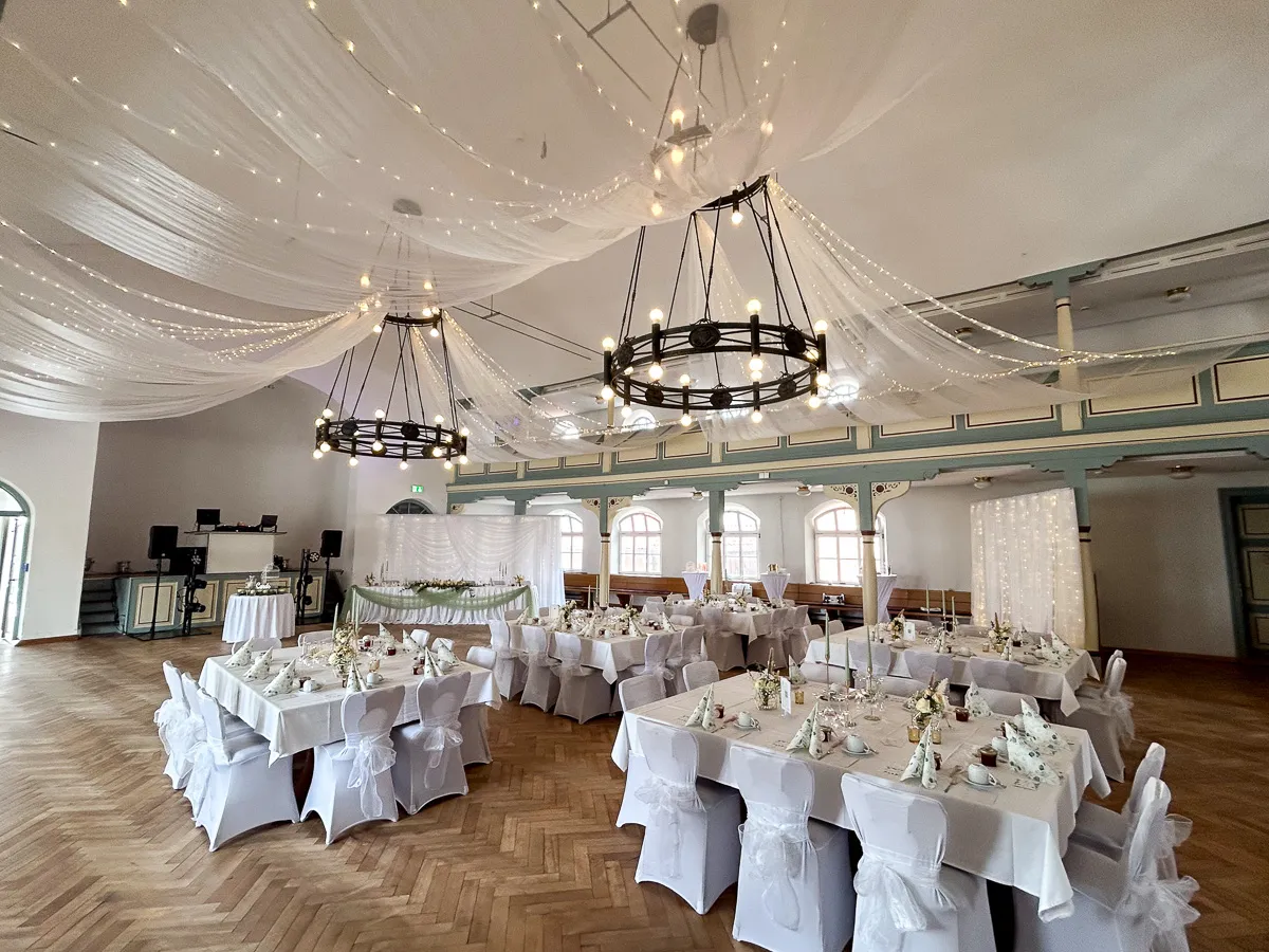 Großzügiger Hochzeitssaal mit mit weißen Stoffbahnen und Lichterketten dekorierter Decke, runden Leuchtern und festlich gedeckten Tischen mit weißen Stuhlhussen.