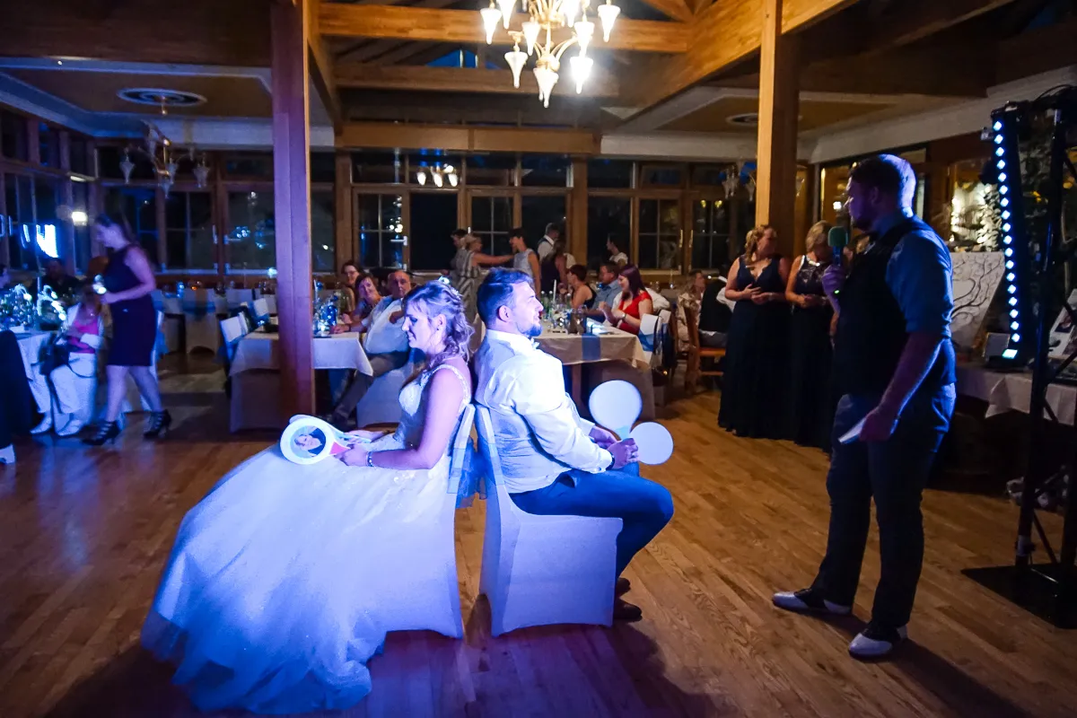 Braut und Bräutigam sitzen Rücken an Rücken und spielen ein Paddel‑Spiel bei der Feier, während ein Moderator mit Mikrofon spricht und Gäste an den Tischen zuschauen.