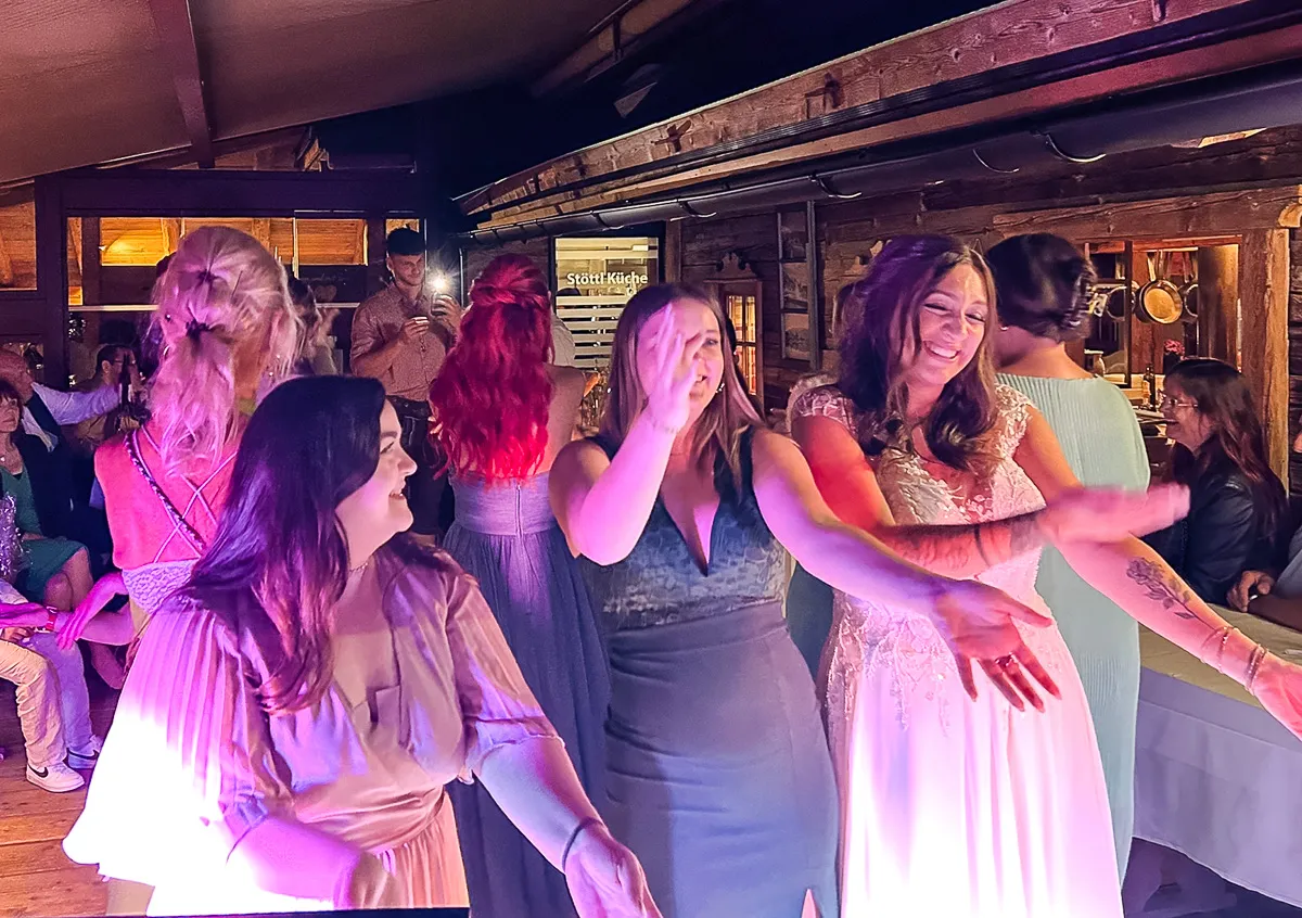 Frauen, darunter eine Braut, tanzen und lachen gemeinsam bei einer Feier in einem rustikalen Raum, der von farbigen Lichtern erhellt wird.