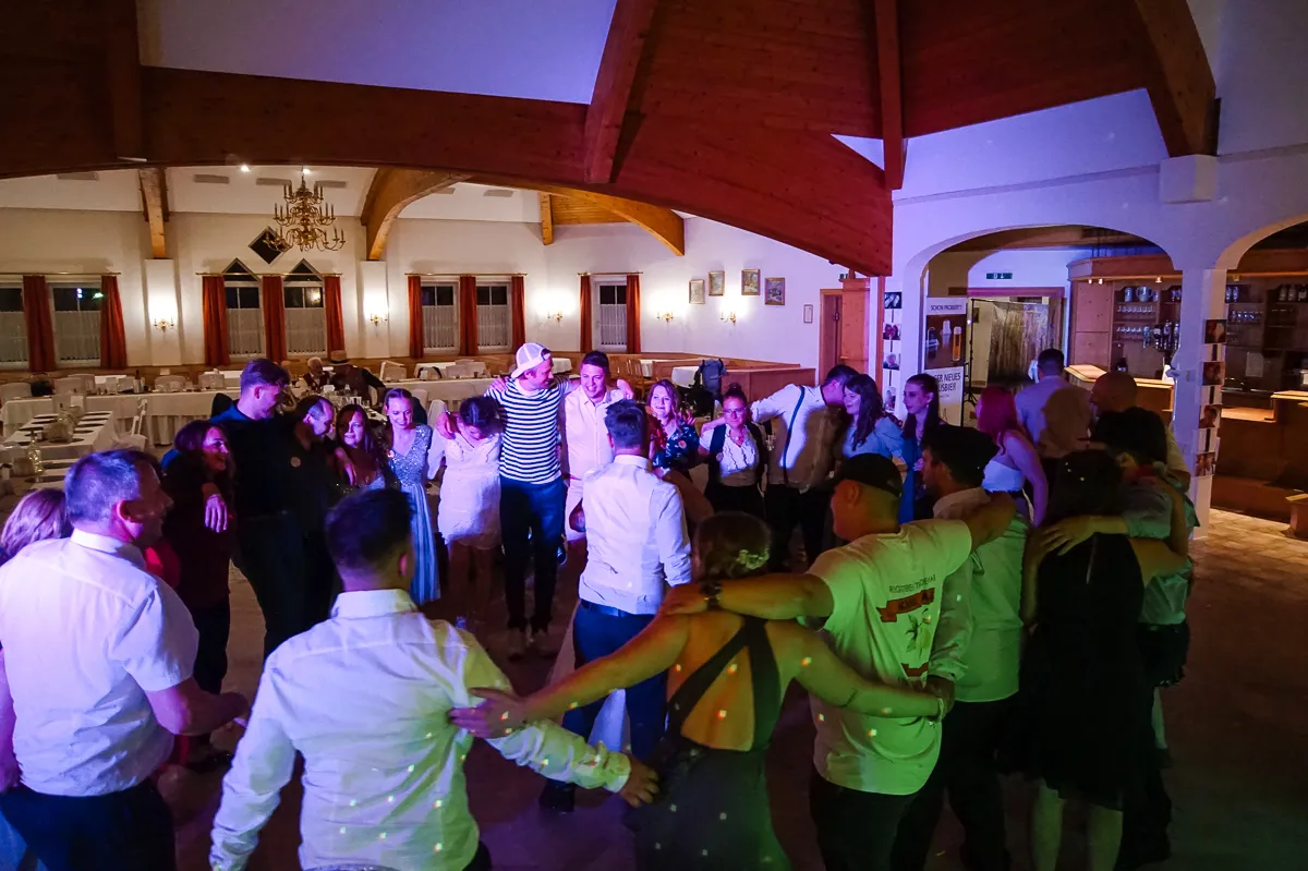 Eine Gruppe von Gästen steht Arm in Arm in einem Kreis auf der Tanzfläche, beleuchtet von bunten Partylampen, in einem großen Festsaal mit Holzbalken.