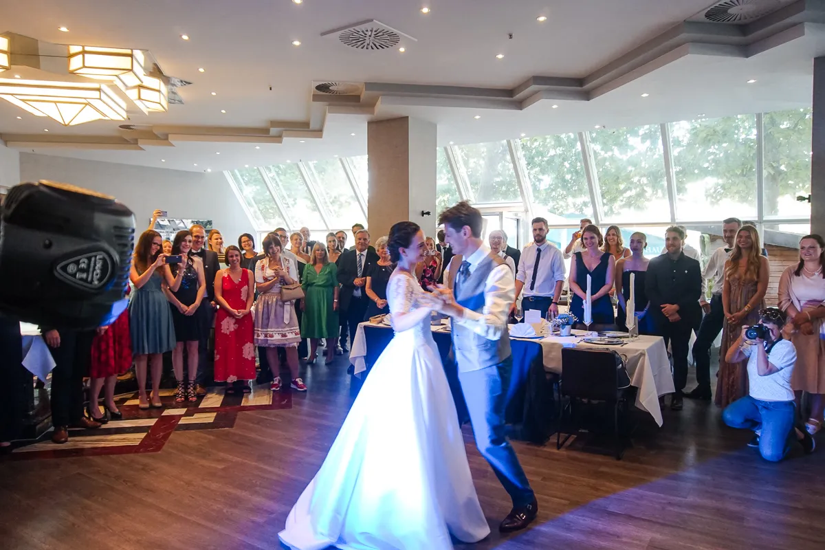 Braut und Bräutigam tanzen in einem hellen Saal mit großen Fensterfronten; zahlreiche Gäste beobachten und fotografieren den Moment.