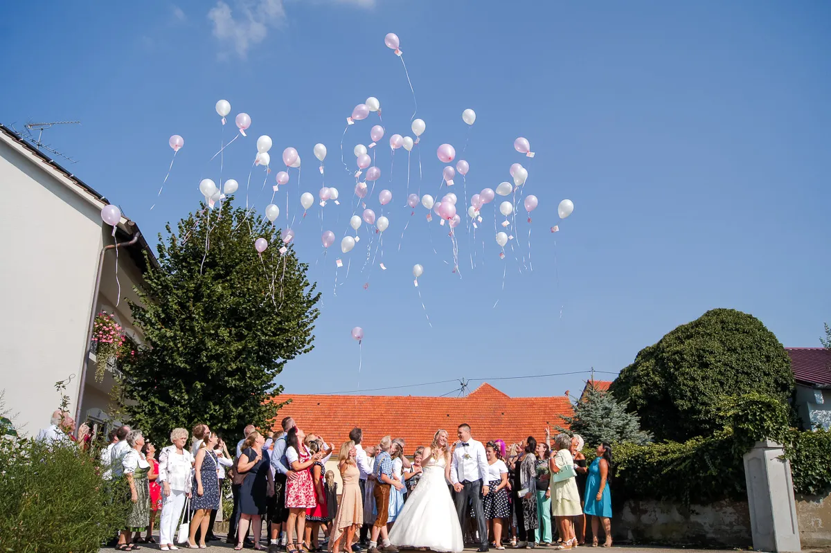 Hochzeitssociety lässt rosa und weiße Luftballons in den blauen Himmel steigen; Braut und Bräutigam stehen Hand in Hand und beobachten.