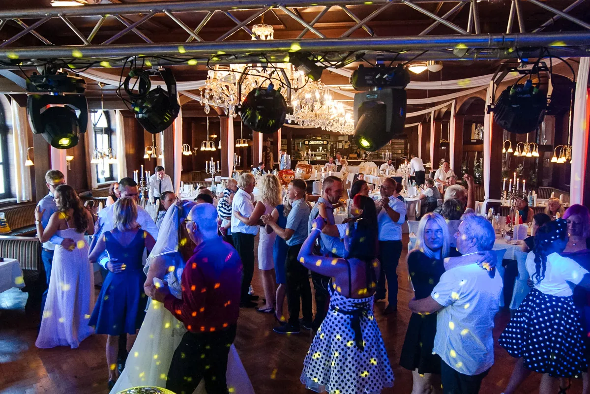 Paare tanzen auf der Tanzfläche eines Hochzeitssaals unter Kronleuchtern und Scheinwerfern, bunte Lichtpunkte reflektieren auf ihnen.