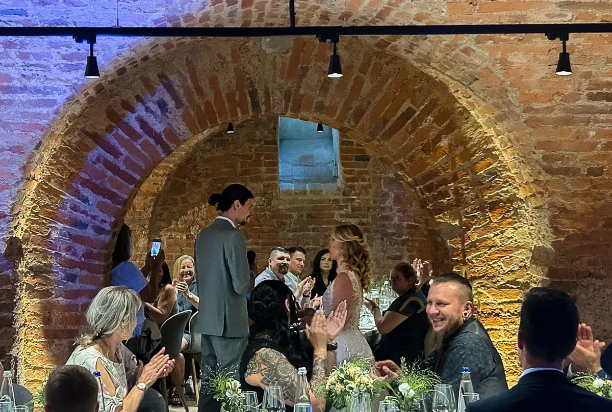 Die Braut und der Bräutigam stehen einander gegenüber in einem historischen Gewölbekeller mit Backsteinbögen, während Gäste an runden Tischen applaudieren.