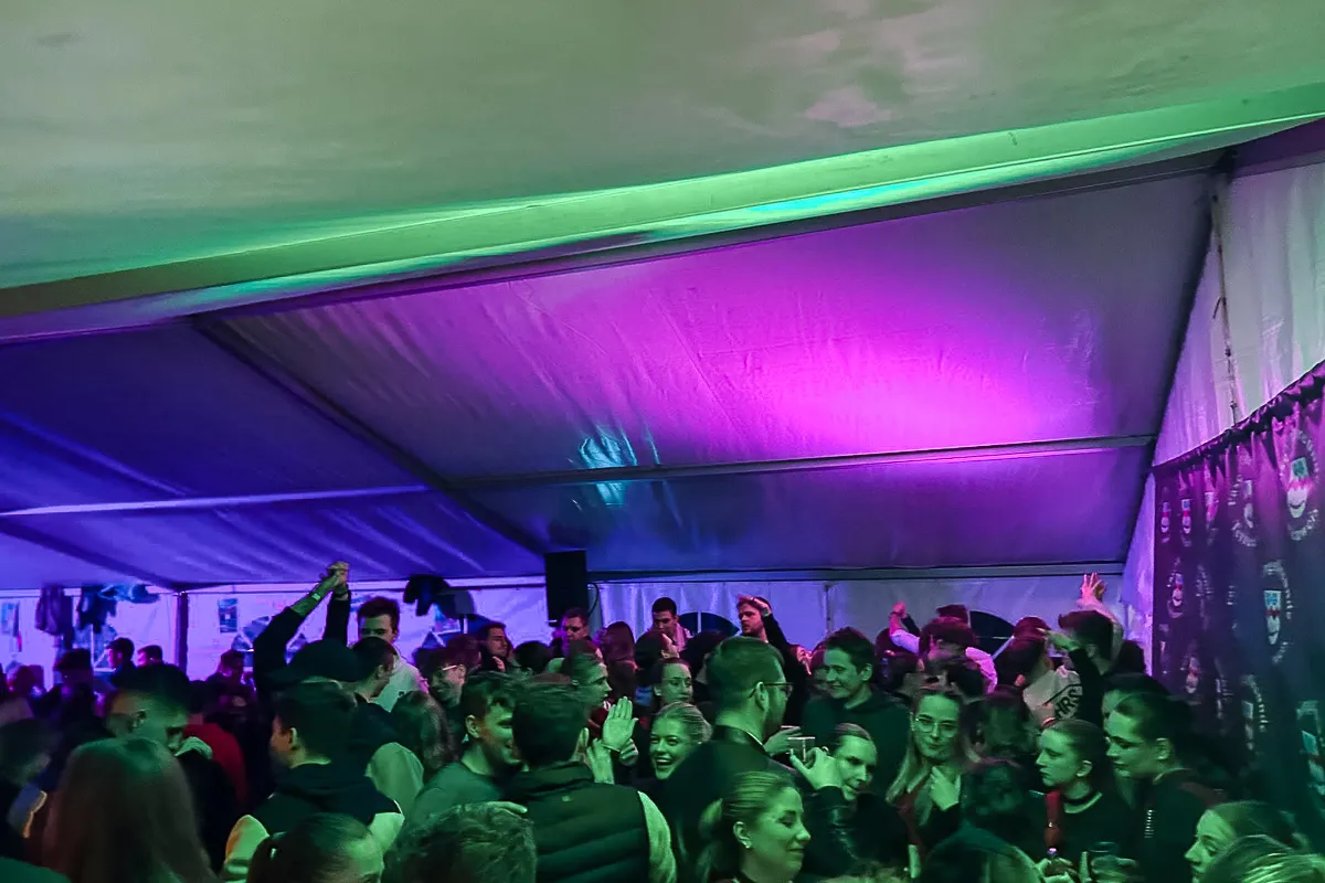 Menschen feiern in einem belebten Partyzelt unter grün‑ und violettfarbigen Lichtern.