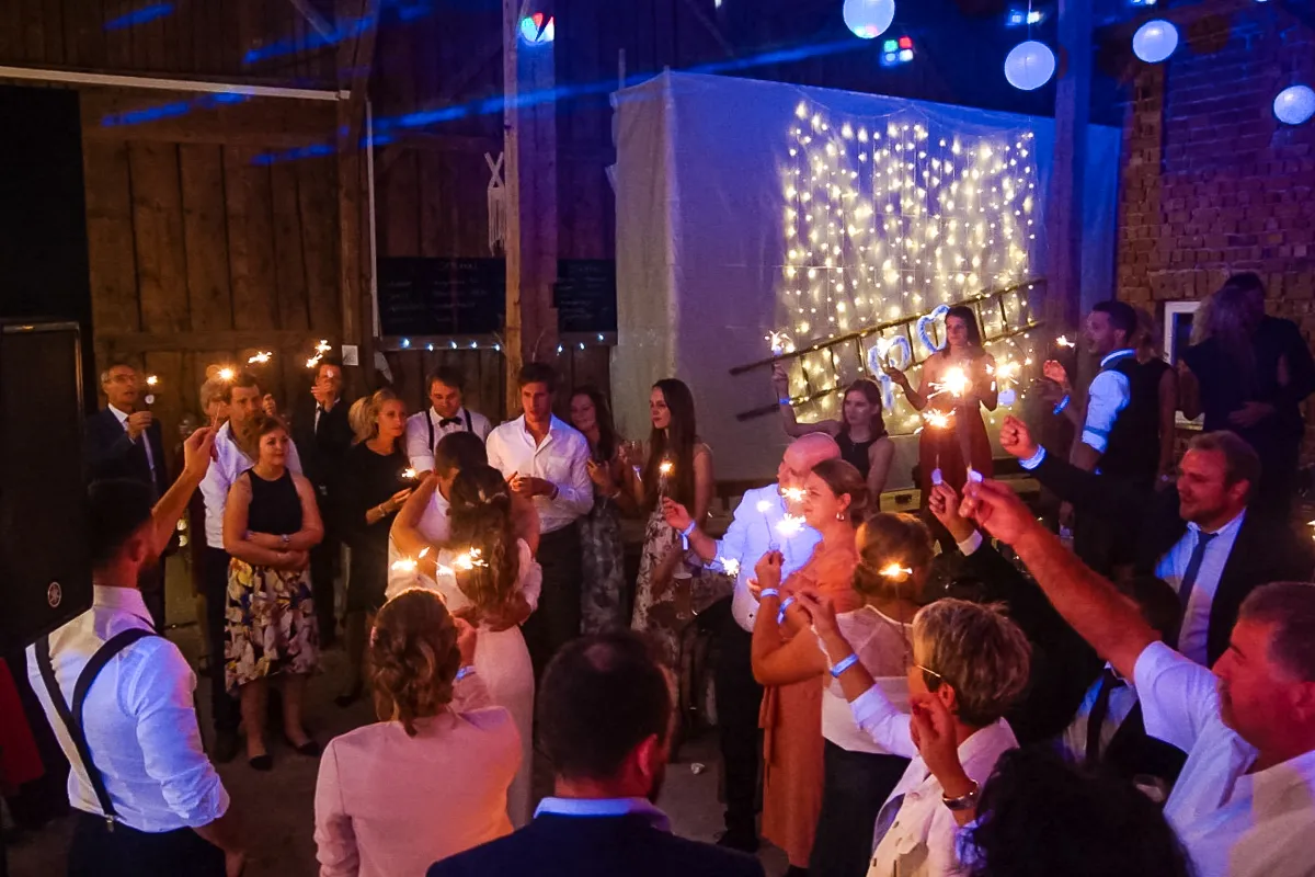 Hochzeitsgäste halten Wunderkerzen hoch in einer rustikalen Scheune, während sie einen feierlichen Moment mit dem Brautpaar erleben, im Hintergrund eine Lichterkette.