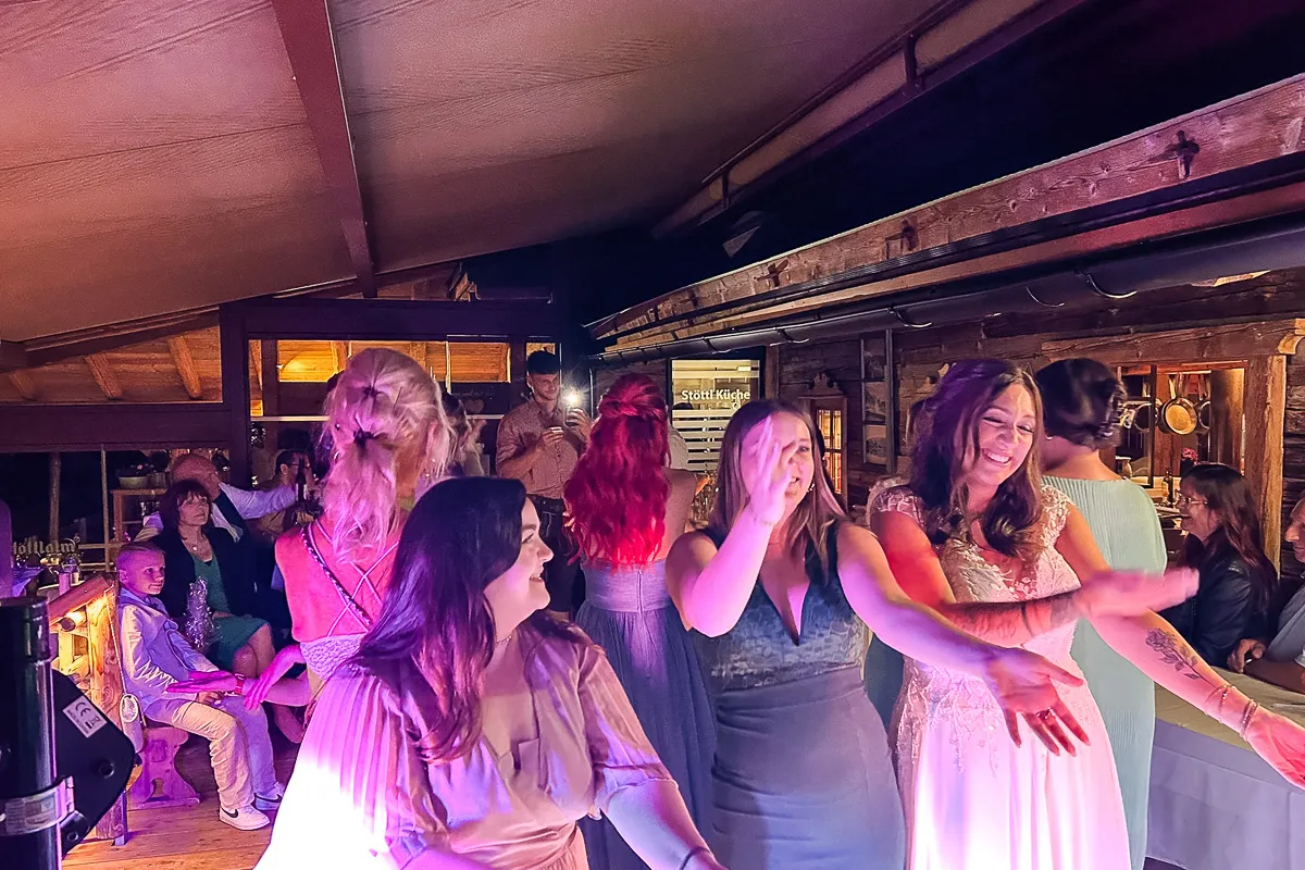 Mehrere Frauen, darunter eine Braut, tanzen und lachen in einer rustikalen Location; warmes Licht fällt auf die Gruppe, während weitere Gäste gemütlich im Hintergrund sitzen.