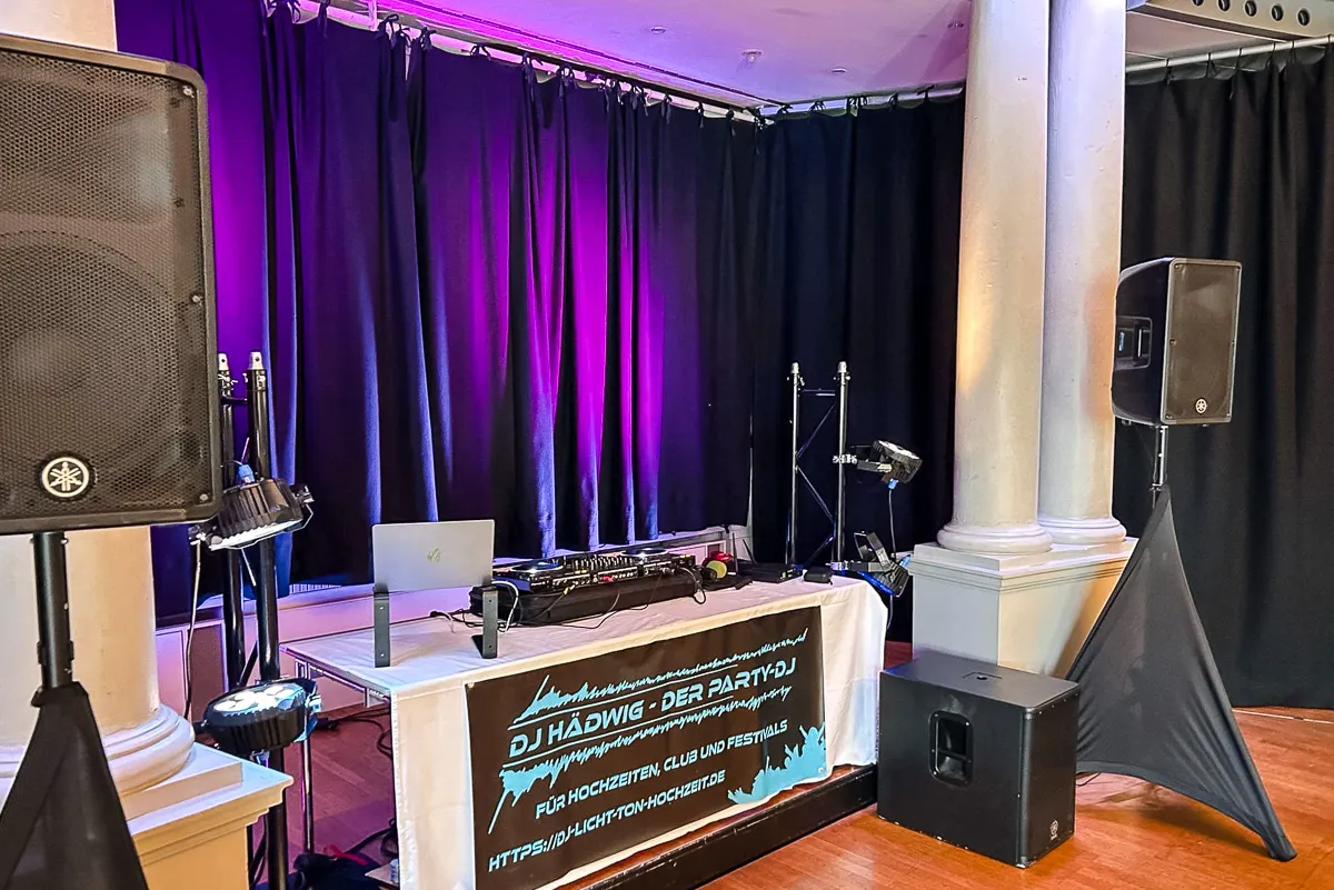 DJ‑Setup mit Laptop, Mischpult und Lautsprechern zwischen Säulen; lilafarbenes Licht beleuchtet den schwarzen Vorhang, und ein Banner mit der Aufschrift „DJ Hädwig – Der Party‑DJ“ hängt vorn.
