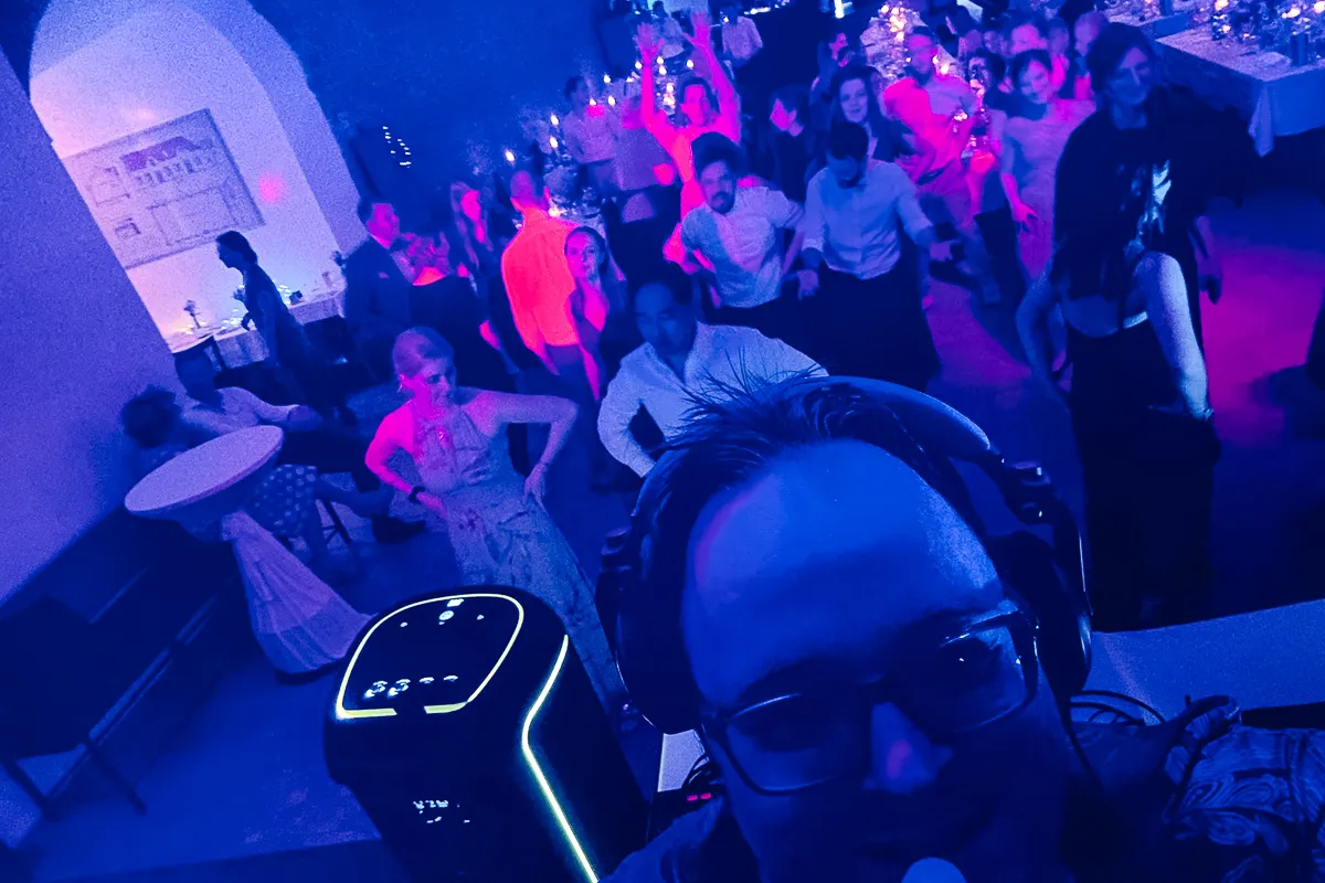 DJ-Pult mit Blick auf die Tanzfläche – im Vordergrund ein DJ-Selfie, im Hintergrund tanzende Gäste im blauen und pinken Licht.