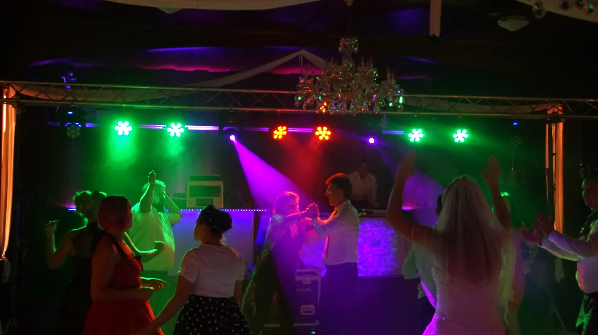 Feiernde Hochzeitsgesellschaft auf der Tanzfläche mit DJ, bunte Beleuchtung in Blau und Pink.