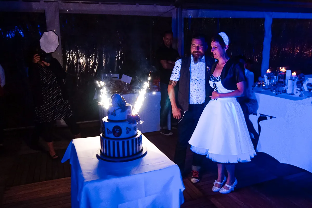 Brautpaar mit Hochzeitstorte und Wunderkerzen – Die Braut im weißen Kleid mit schwarzem Bolero und der Bräutigam mit Weste lächeln neben einer festlich dekorierten Torte mit Fontänen.