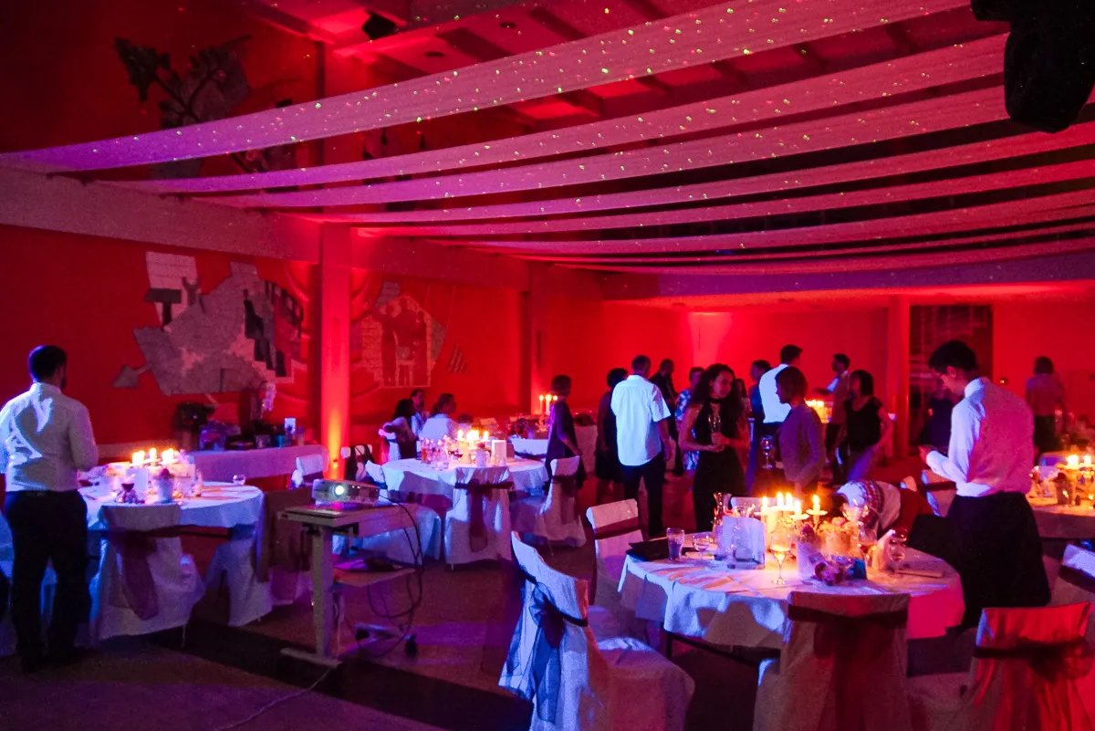 Hochzeitsfeier mit roter Lichtstimmung – Gäste sitzen und tanzen in einem festlich dekorierten Saal mit roten Wänden und Kerzenlicht.