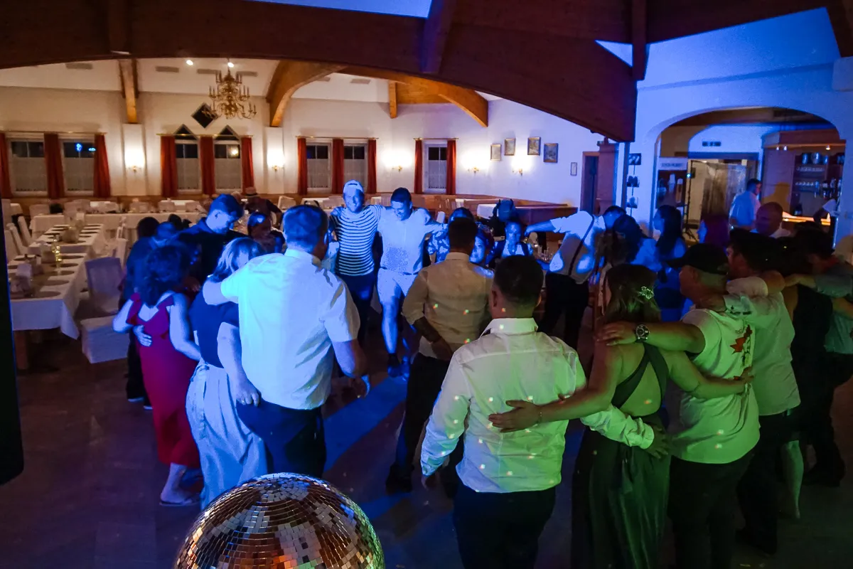Gäste tanzen im Kreis bei Hochzeitsfeier – Menschen stehen Arm in Arm auf der Tanzfläche, im Vordergrund eine Discokugel, im Hintergrund leere Tische.