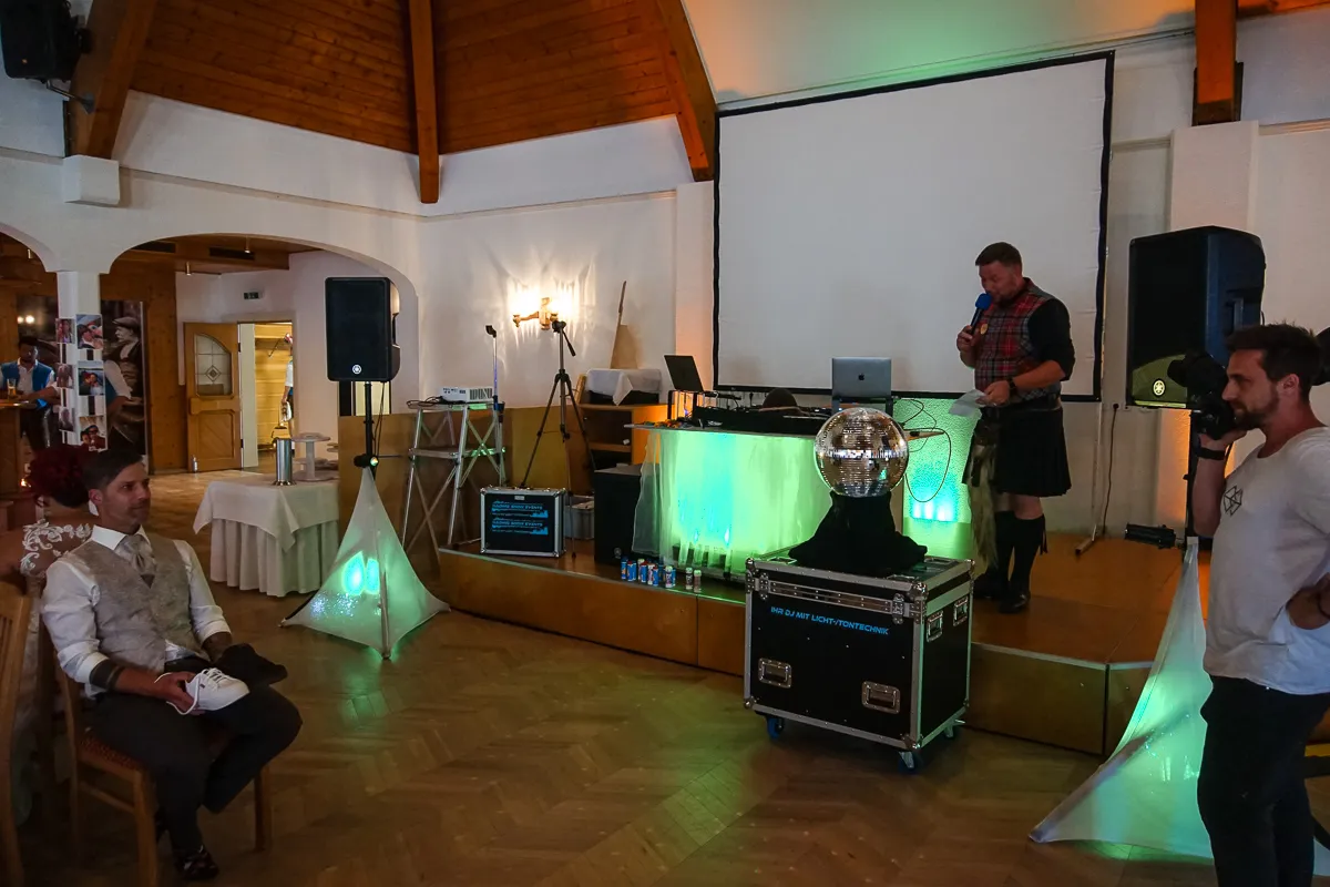 DJ-Setup mit Lichttechnik – Ein DJ-Pult mit Laptop, Lautsprechern, Beleuchtung und Discokugel, daneben ein Moderator im Kilt mit Mikrofon.