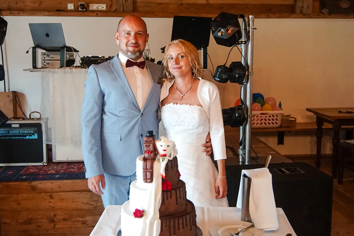 Brautpaar posiert neben einer Hochzeitstorte mit witzigen Figuren, im Hintergrund DJ-Technik und Lichtanlage.