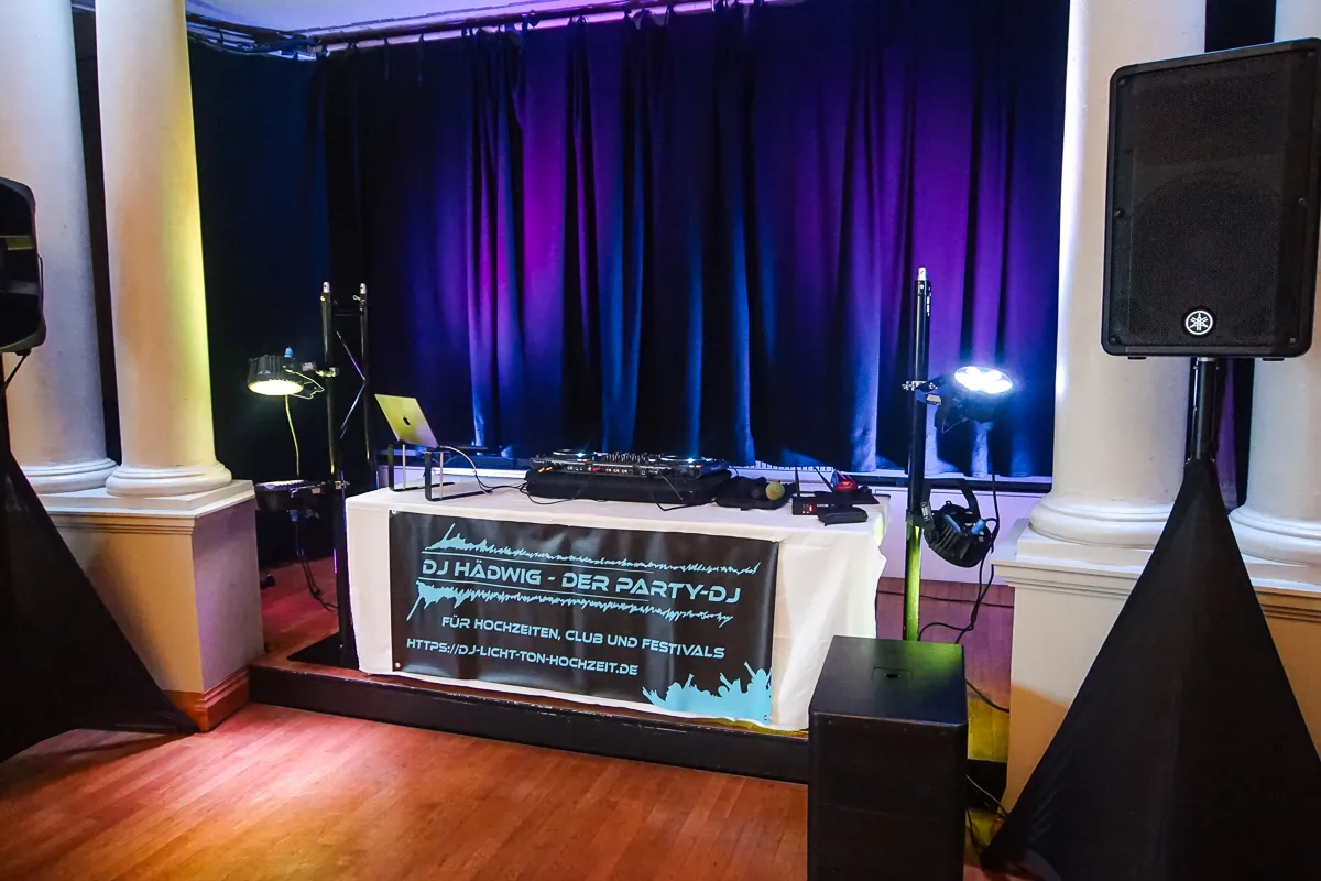 Modernes DJ-Pult mit Banner „DJ Hädwigs – Der Party-DJ“ vor schwarzem Vorhang und farbiger Lichtbeleuchtung.