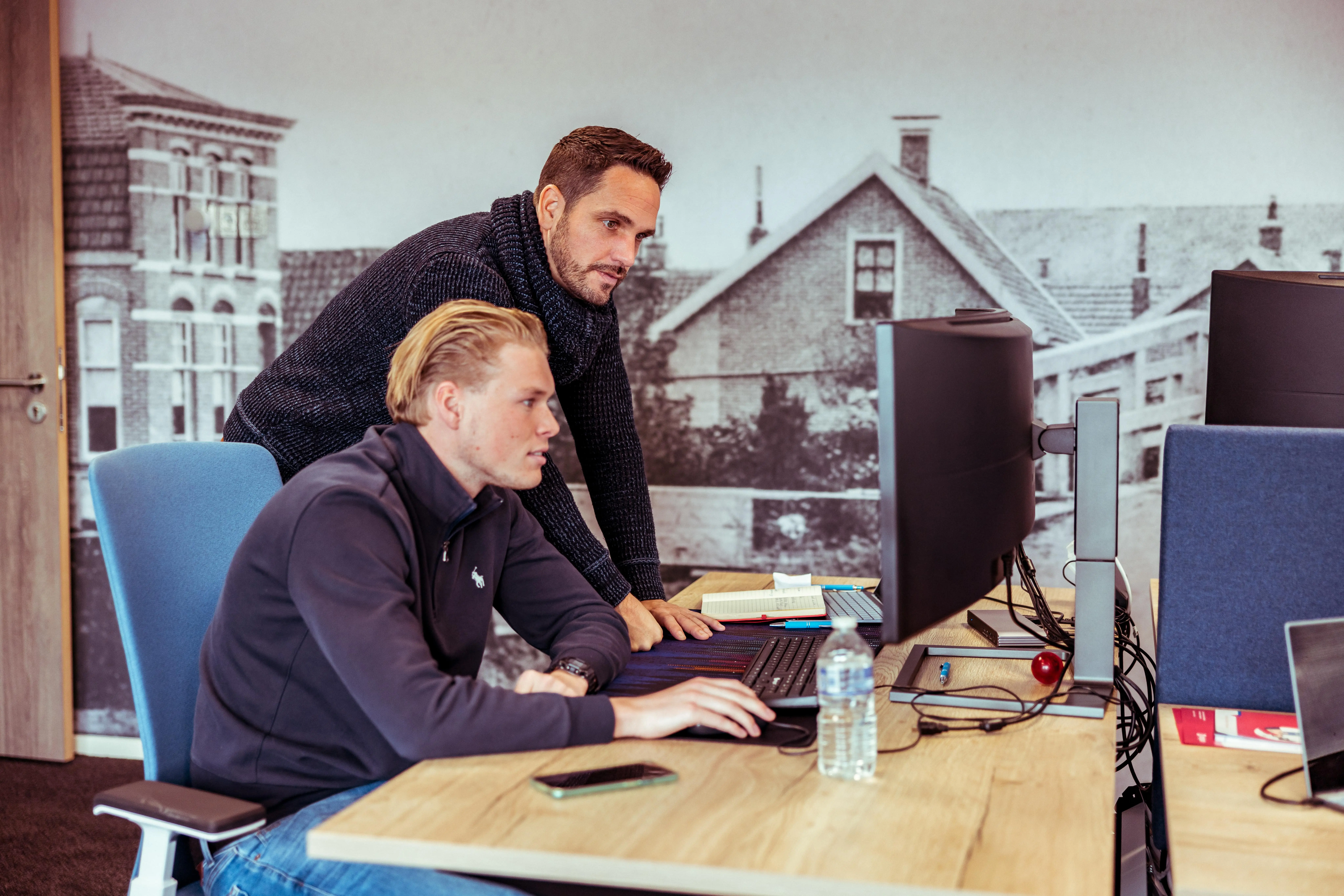Two men working together at a desk with a computer monitor, notebook, and water bottle in a modern office.