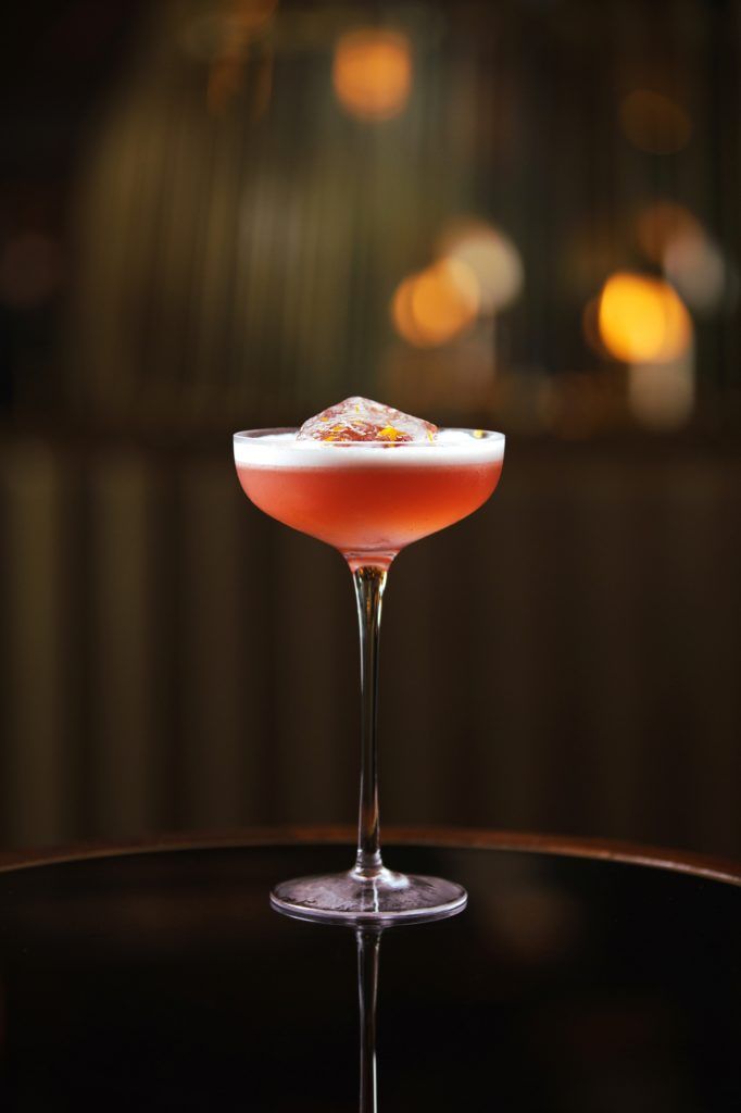 Elegant pink cocktail in a coupe glass with a large ice cube and orange zest, set against a dimly lit background.
