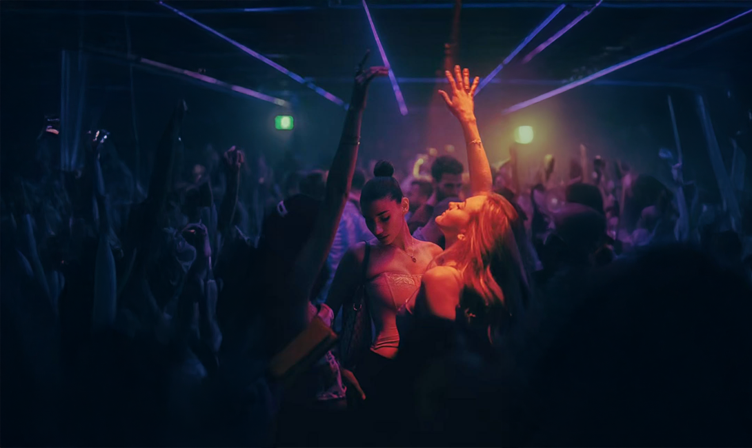 Crowd dancing energetically with raised hands in a dimly lit nightclub with purple and orange lighting.