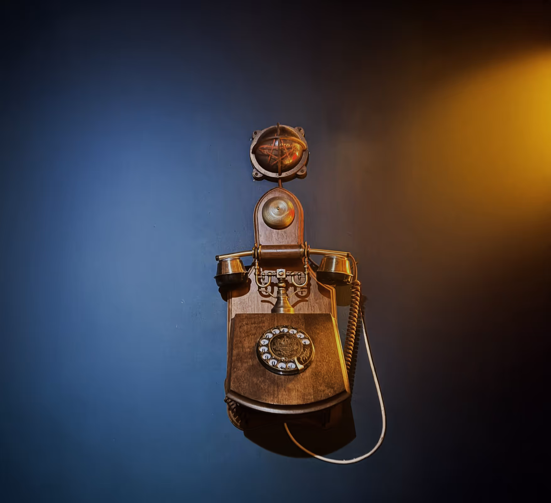 Vintage wooden wall-mounted rotary phone with brass accents on a dark blue wall with warm light.