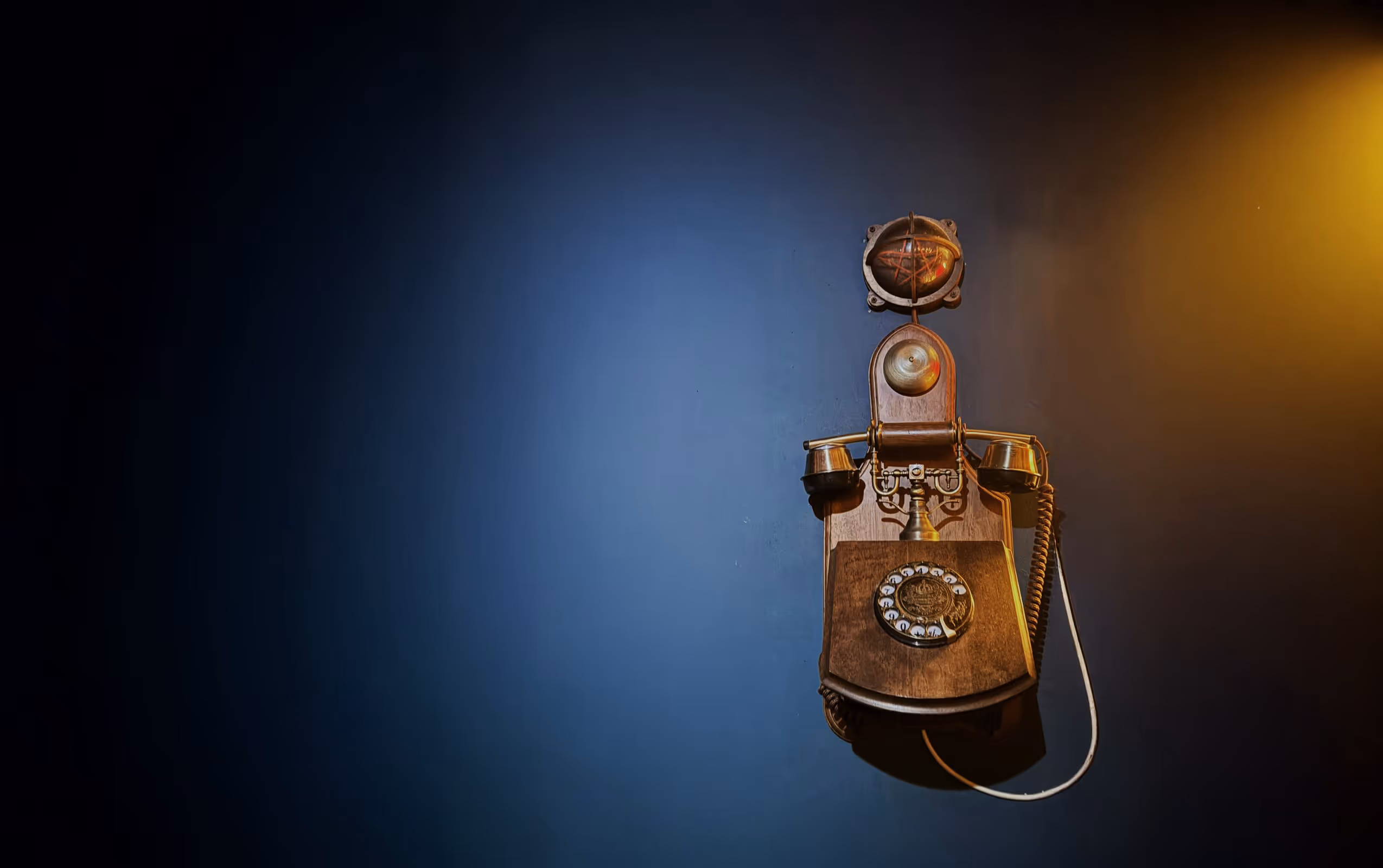 Vintage wooden rotary wall telephone mounted on a dark blue wall with warm light on the right.