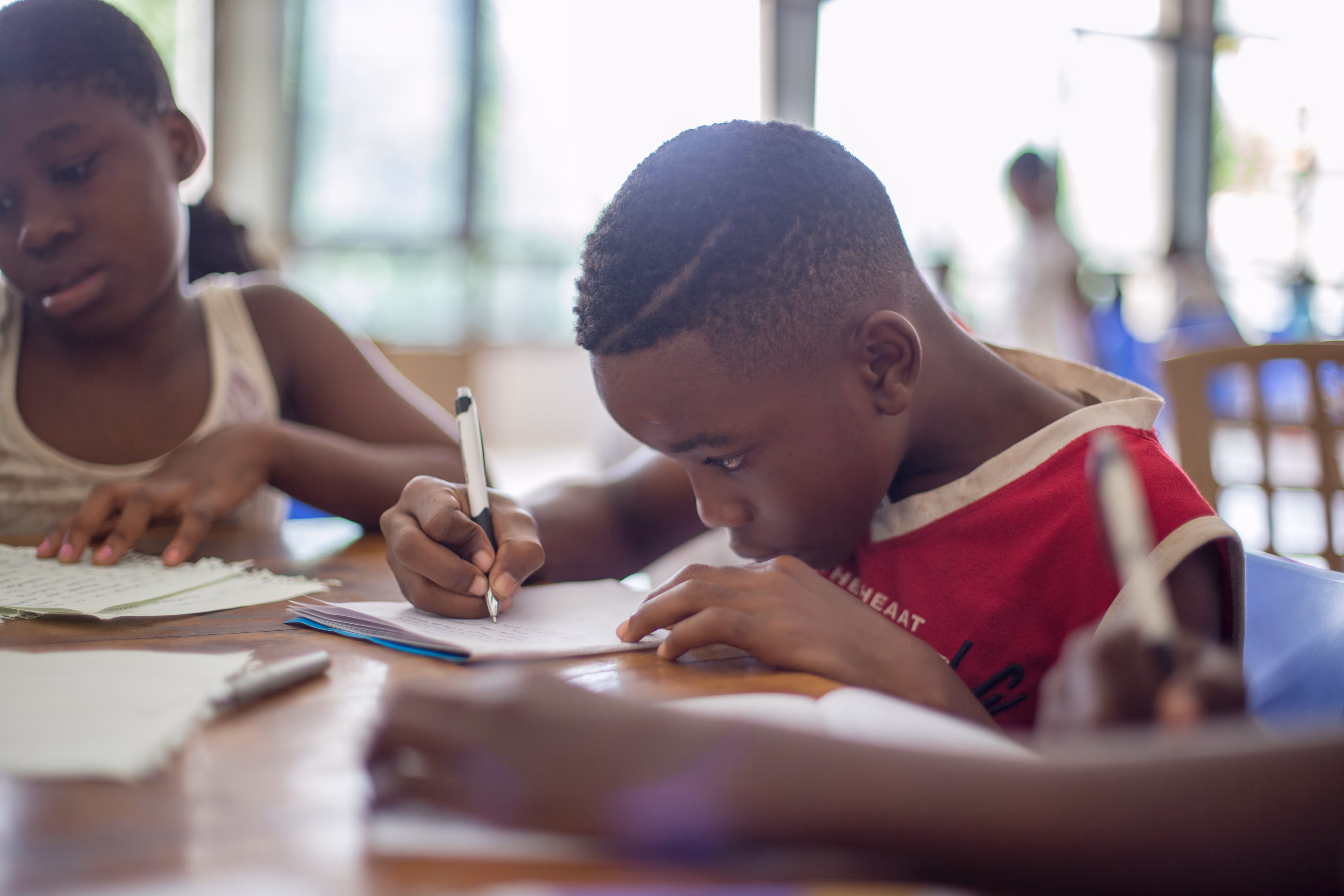 Two children sitting in chairs writing on paper at a desk